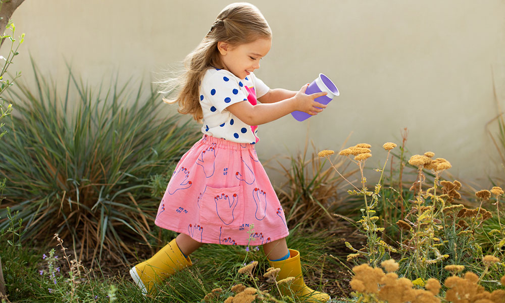 A young girl runs through a garden holding a purple Munchkin Miracle 360° sippy cup