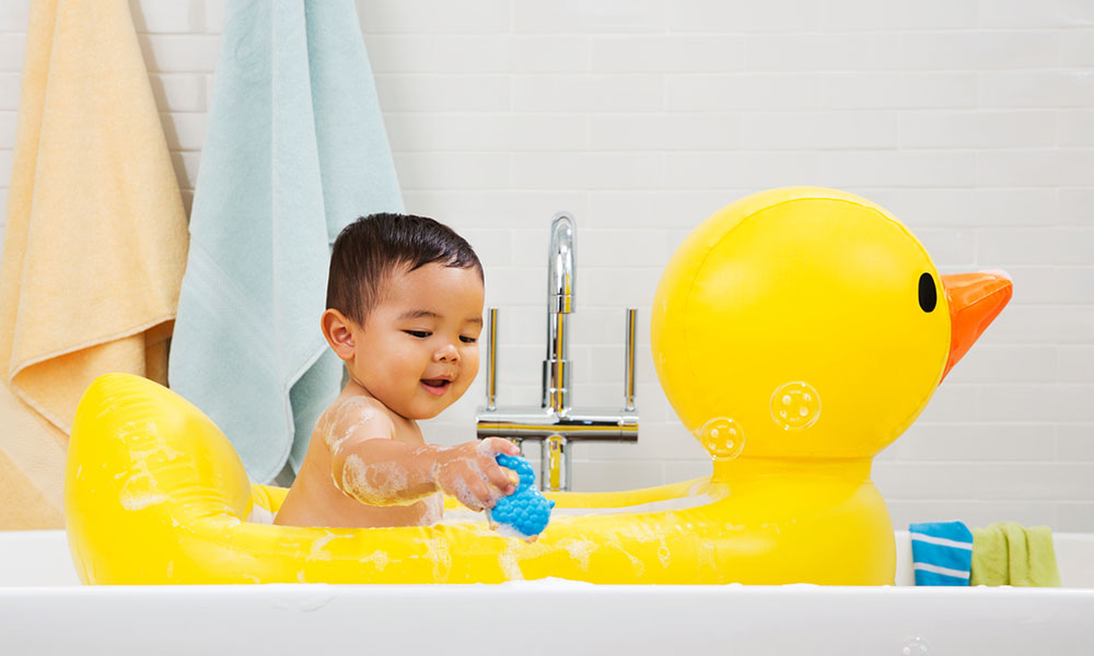 A baby boy sits in a yellow Munchkin inflatable duck bath tub and plays with a bath toy.