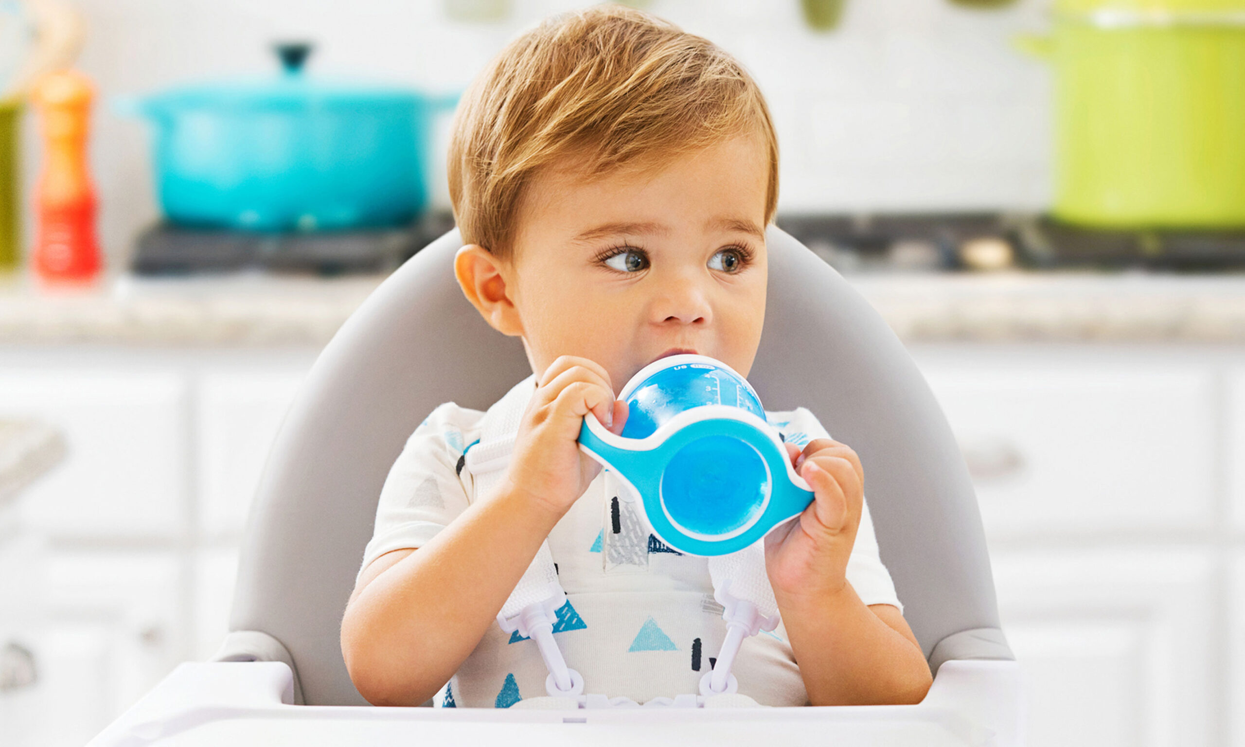 Young boy sits in a high chair in a kitchen, drinking from a blue Munchkin Gentle Transition Cup