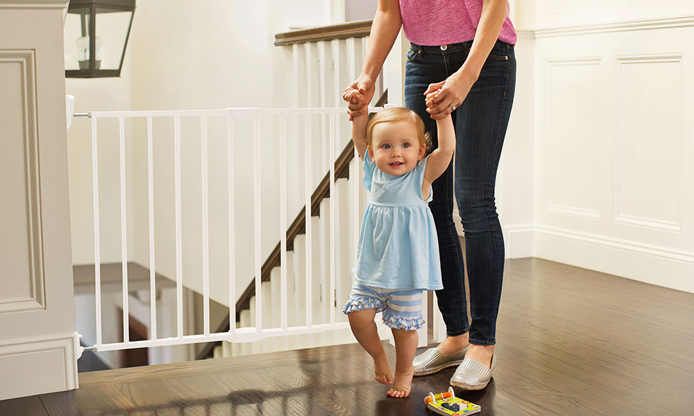 A mum standing in front of a Munchkin safety gate helps a toddler learning to walk