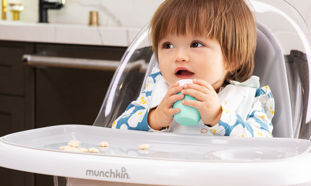 A toddler sits in a highchair holding a mint colour Munchkin C'est Silicone 2oz/118ml Open Cup.