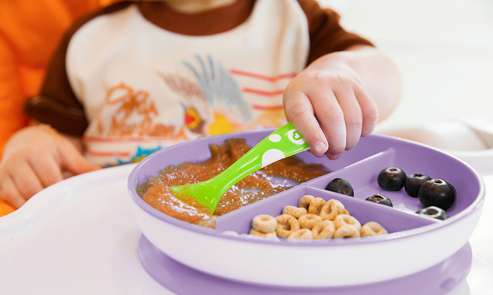 Close up of a purple Munchkin Divider Suction Plate on a highchair, and a toddler's hand picking up food with a spoon