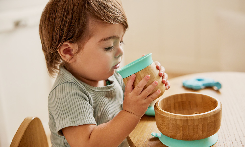 Child drinking from a bamboo open cup with a bamboo suction bowl on the table.