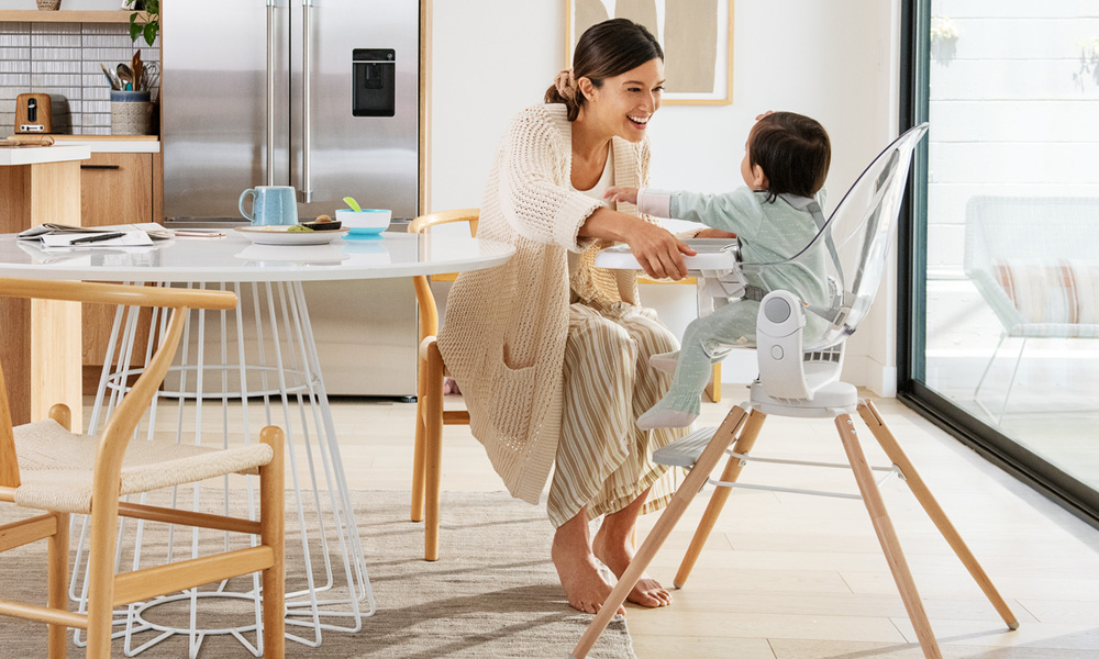 Mother sat next to a baby sitting in a highchair. Women is smiling at the baby.