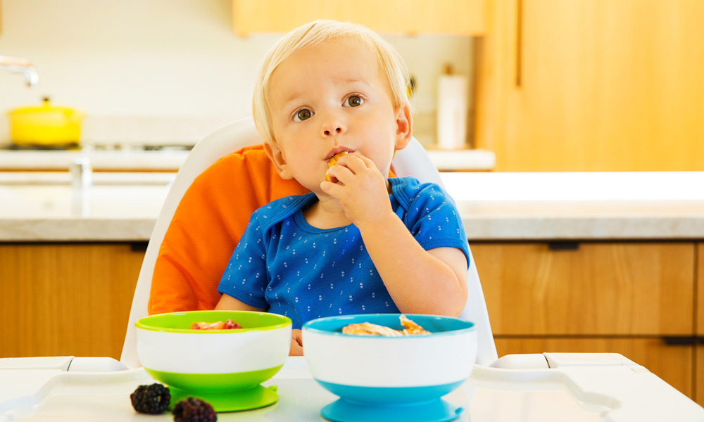 Boy sat in highchair eating out of suction bowls.