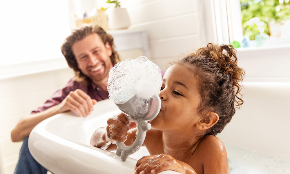 Girl blowing bubbles through the Munchkin Bubble Bestie. Man watching and laughing.